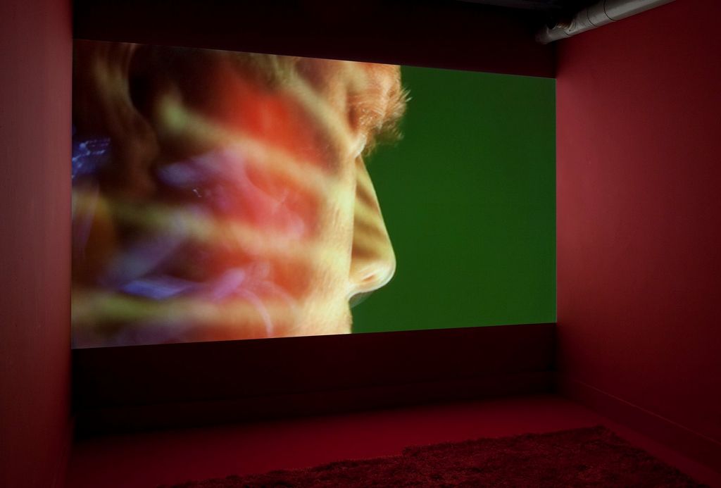 Installation view of the work "The Quickening" by artist Sue de Beer with walls painted burgundy. The still shows a close-up of a human face, seen from the side. Sue de Beer, Sammlung Goetz Munich