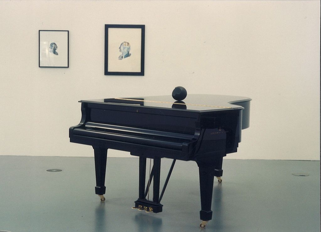 This photograph depicts a black concert grand piano on which a small, likewise black and oval glass sculpture is lying. On the white wall behind it are collages, which are silhouettes of the profile of past American presidents.