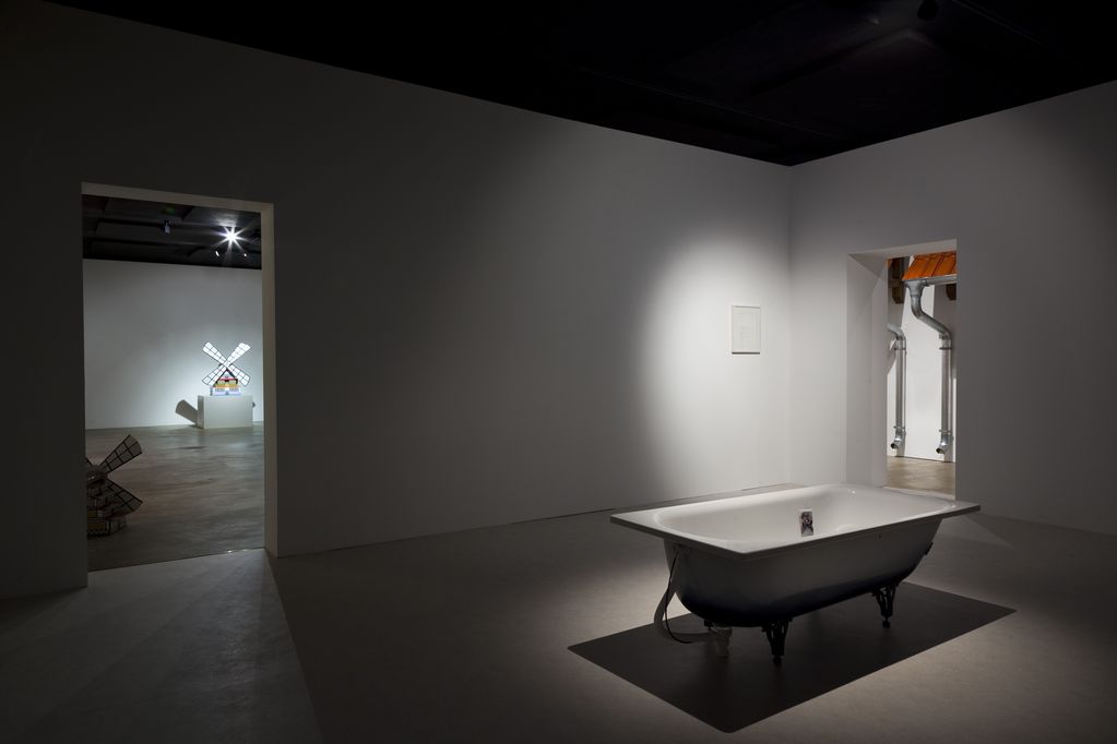 Exhibition view showing a single white bathtub in the center of a white room from which two rooms lead off. The room on the left reveals miniature windmills, while the room on the right gives a view of a silver rain gutter complete with a red tiled roof. Andreas Slominski, Sammlung Goetz Munich