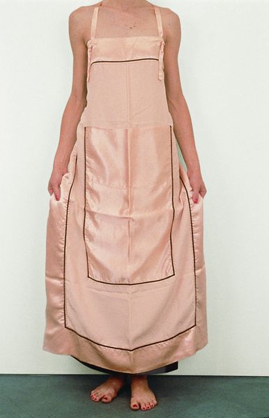 This picture shows the body of a slender woman wearing a light pink apron dress by the artist Andrea Zittel. The apron dress can also be converted into a wall object and is made of silk.