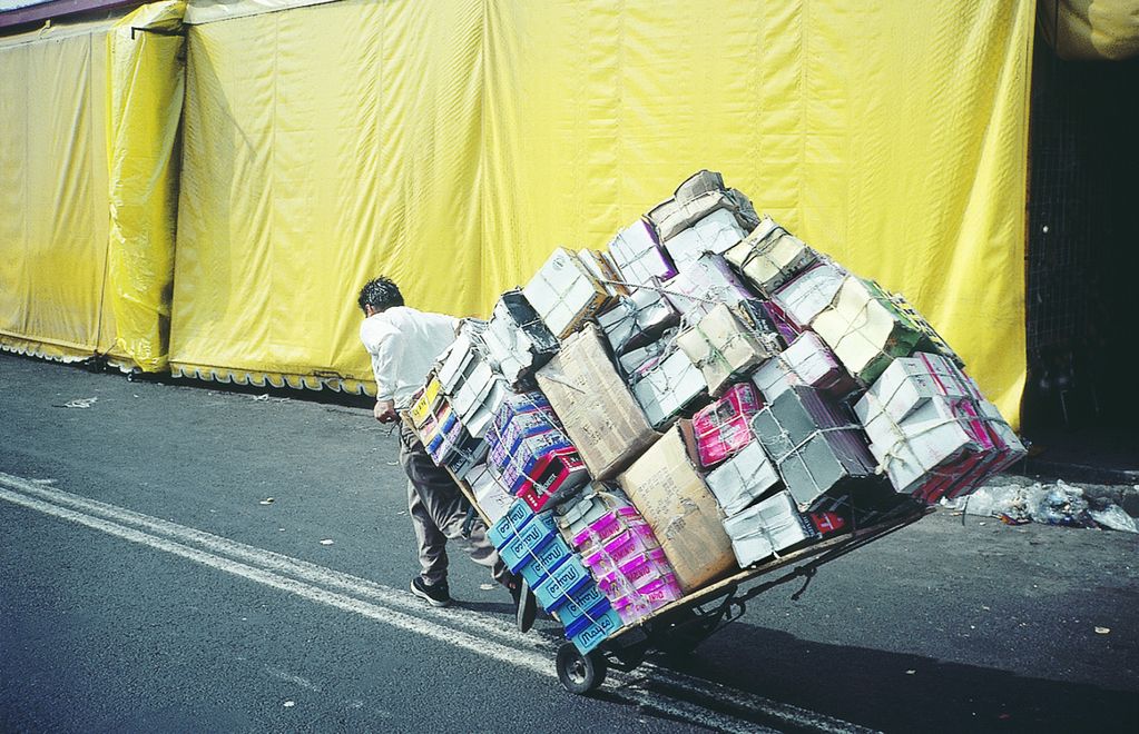 Video Still, welches einen schwer tragenden Mann zeigt, der auf einer geteerten Straße eine Menge Kartons auf einer Sackkarre hinter sich herzieht. Francis Alÿs, Sammlung Goetz München
