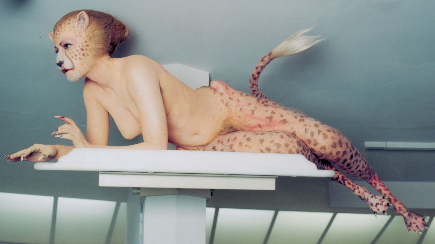 This photograph shows a half-lying leopard human lady, who has the legs, face and tail of a leopard and who otherwise has human attributes. Blood can be seen on her mouth and her false, claw-like fingernails. She is in a brightly lit white room on a pedestal.
