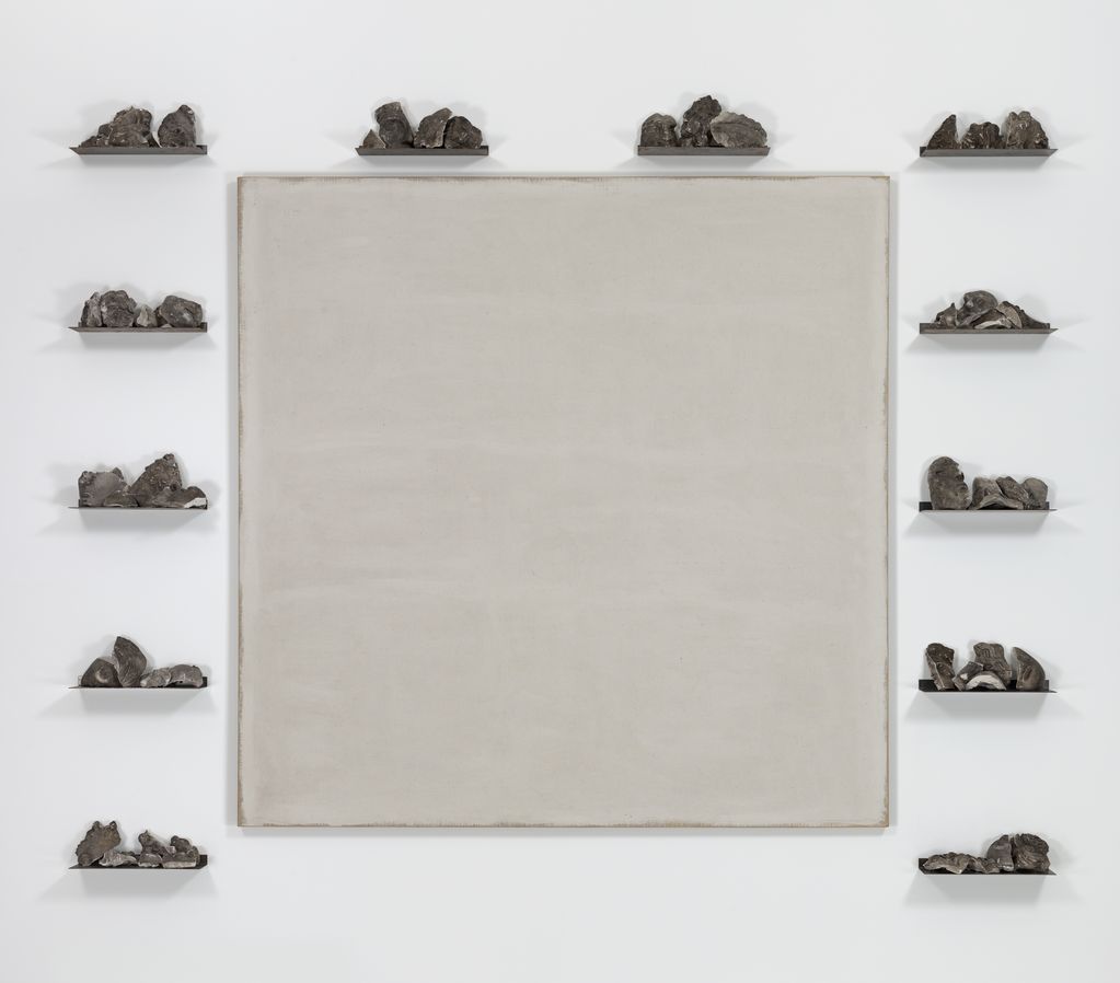 Diese Arbeit von Jannis Kounellis beinhaltet eine quadratische, weiß bemalte Leinwand. Um diese Leinwand herum sind Bruchstücke von Gipsabgüssen antiker Köpfe auf schmalen Ablagen platziert.
