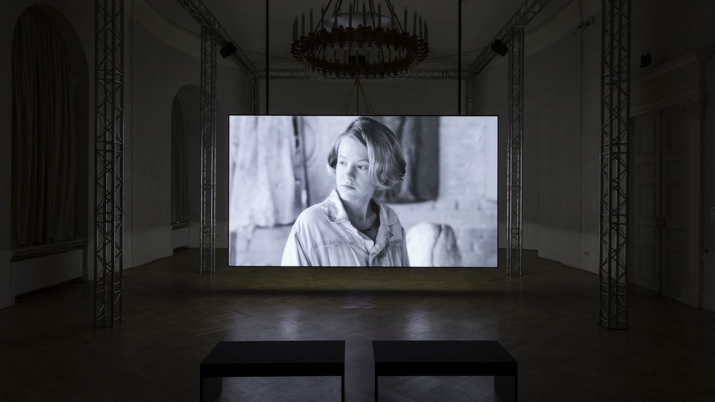 Exhibition space with large screen showing a video still in black and white of a young blonde woman. Teresa Hubbard/Alexander Birchler, Sammlung Goetz Munich