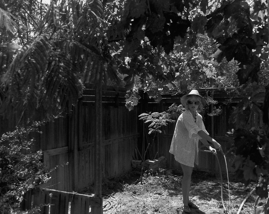 Schwarzweißfotografie von einer Frau, die mit einer hellen Tunika und Sonnenhut bekleidet ist und in die Richtung des Betrachtenden blickt, während sie ihren Garten mit einem Gartenschlauch gießt. Cindy Sherman, Sammlung Goetz München
