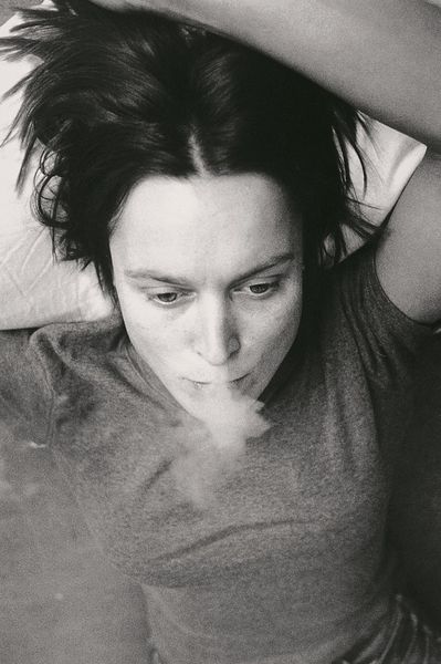 This black and white photograph is a self-portrait of the artist Sarah Lucas seen from above. She is lying in bed smoking a cigarette with one arm above her head.