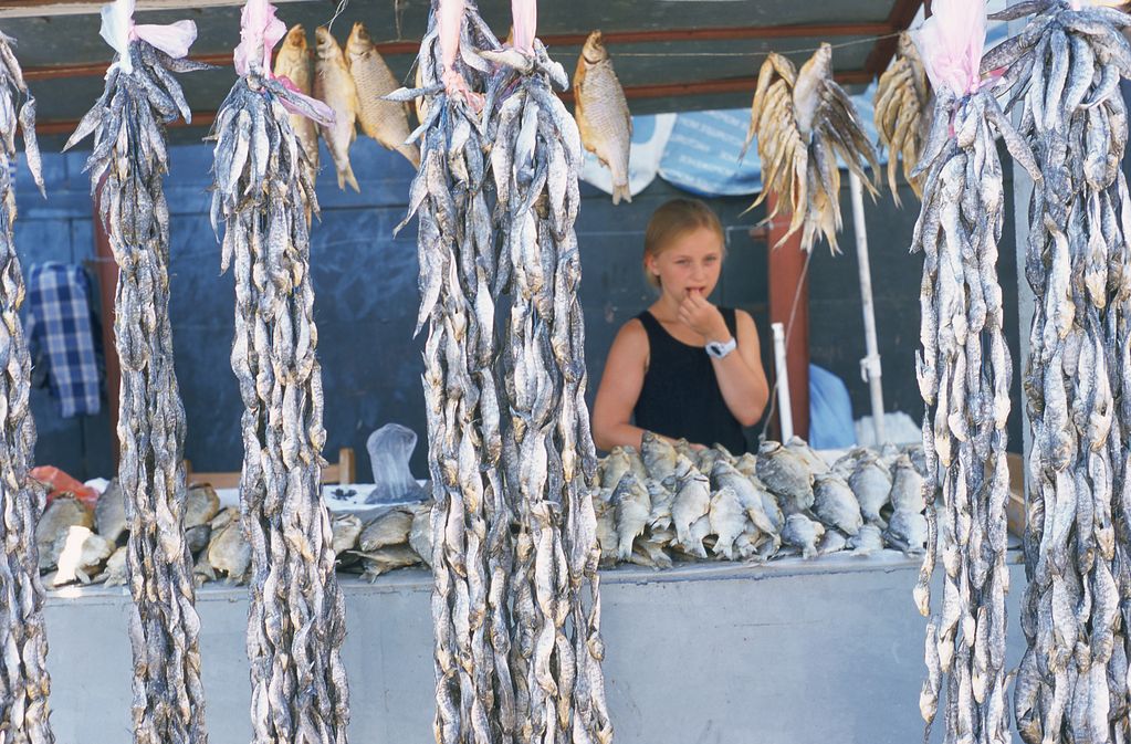 Photograph of a girl standing behind the table of a dried fish stand thoughtfully putting something in her mouth. Ulrike Ottinger, Sammlung Goetz Munich