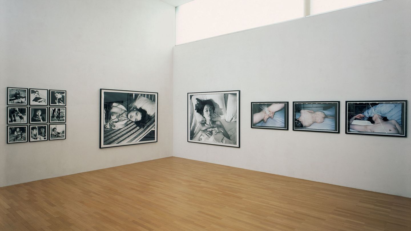 This installation view shows photographic works by the Japanese artist Nobuyoshi Araki. Nine smaller formats in rows of three hang on the left wall, consisting of black and white photographs of frivolous Japanese girls in school uniforms. To the right is a large-format black-and-white photograph of a tied up woman with her upper body exposed. On the right wall is again a large-format black-and-white photograph of a young woman whose head and neck peek out of a bedspread. Next to it hang color photographs of partial views of naked women, whose intimate parts have been scratched away with a sharp object. 
