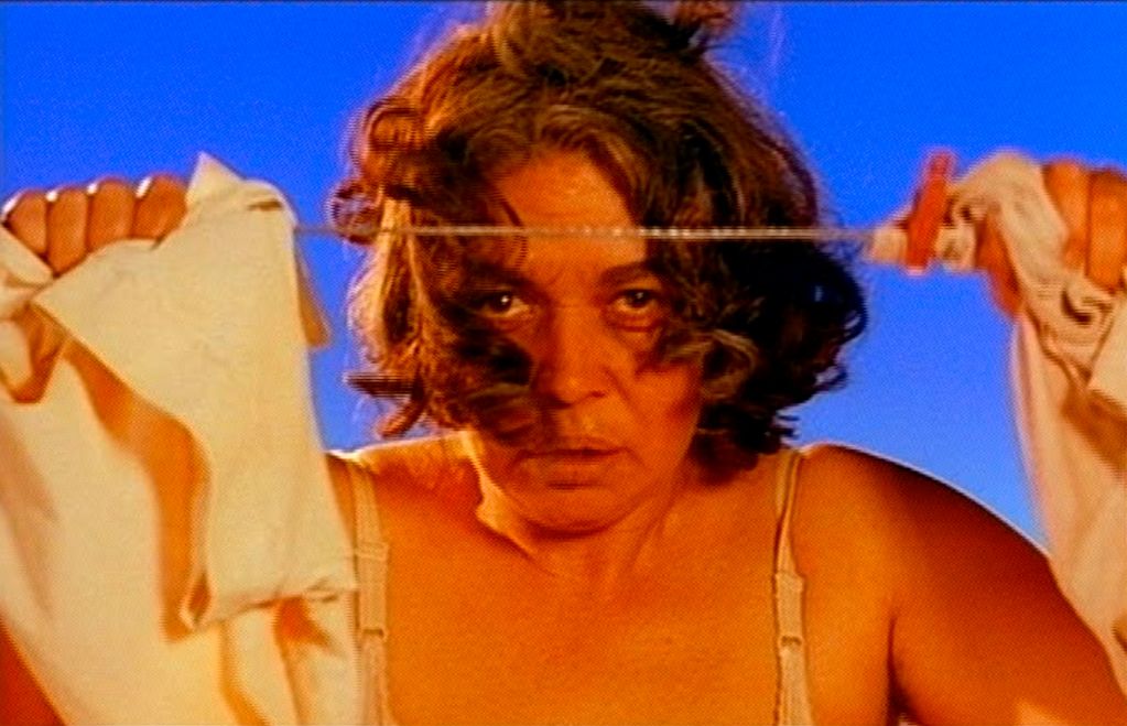In this video still, a middle-aged brunette woman looks between two pieces of clothing hanging on a clothesline. The coloration appears unnaturally saturated.