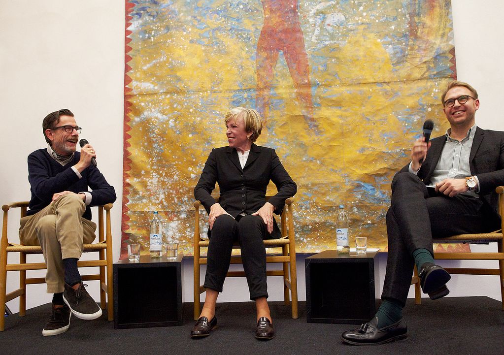 Photograph of a podium on which the artist Marcel Odenbach, the collector Ingvild Goetz and the curator Karsten Löckemann are sitting and having a conversation. Behind them is a large-format painting by Michael Buthe. Sammlung Goetz Munich