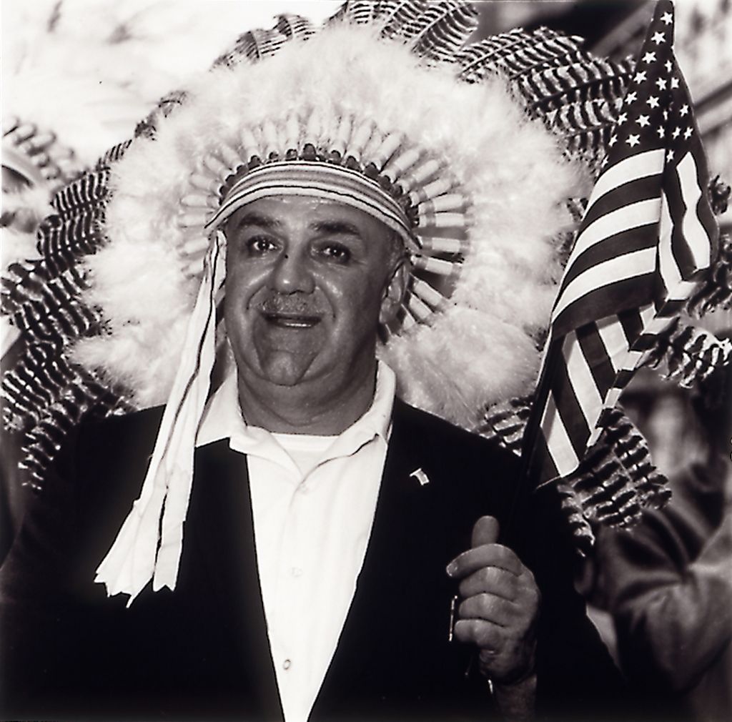 This black and white photograph depicts a man in a white polo shirt, dark jacket and an Indian headdress consisting of feathers.