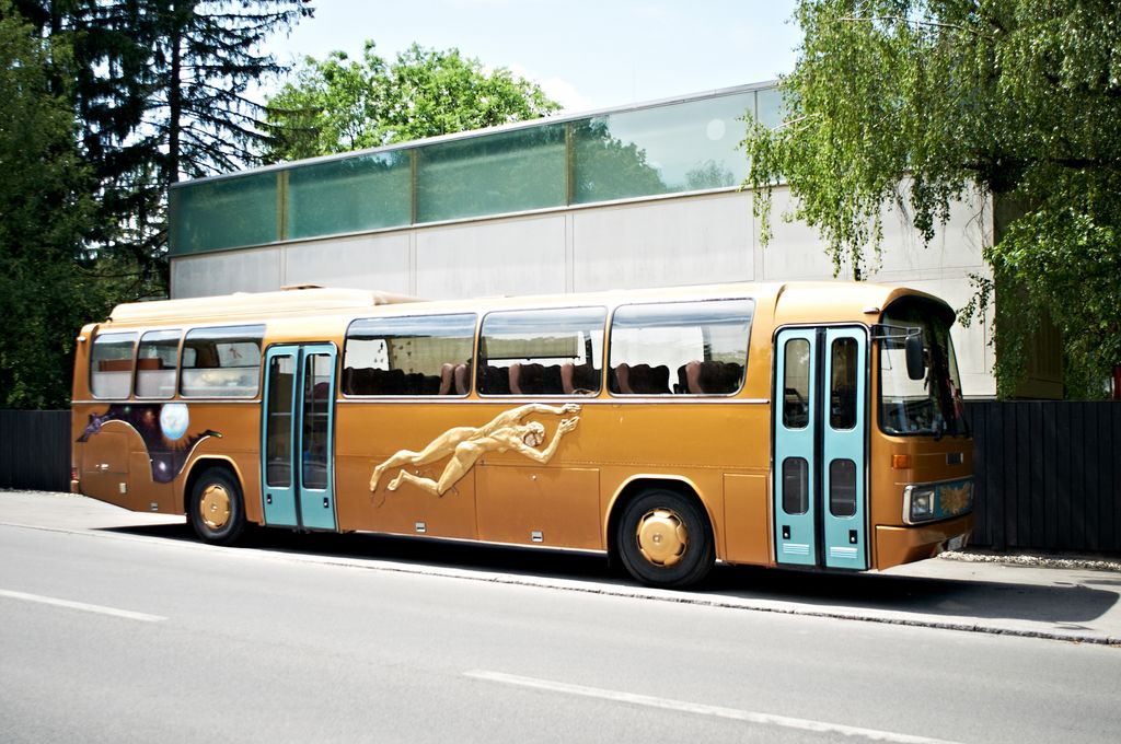 Photo of a golden bus in front of the garden wall of the Sammlung Goetz. The bus is emblazoned with a likewise golden figure of a naked woman in rear view, who appears to be floating. Paweł Althamer, Sammlung Goetz