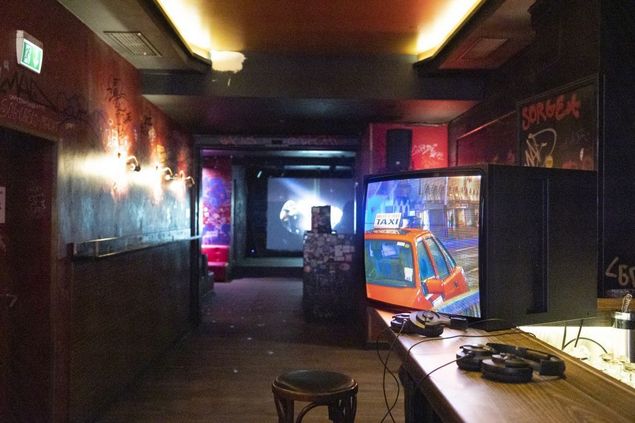 Suite of rooms in a bar/club with red walls covered with graffiti and inscriptions. On the left, a bar counter protrudes into the picture. On it is a monitor with two headphones. A red cab can be seen on the screen. 