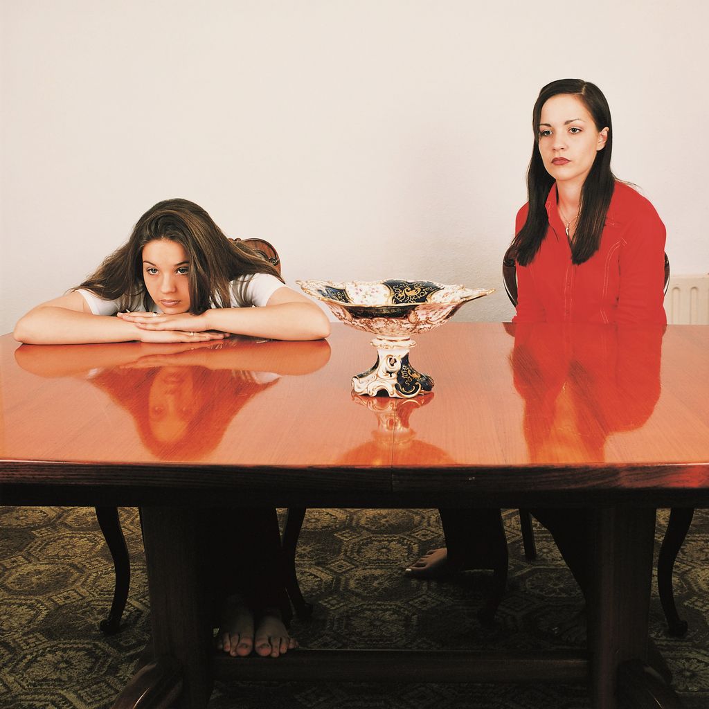 This photograph shows two women sitting at a shiny table. In the middle is a silver fruit goblet. The young women look into the distance, waiting and bored.