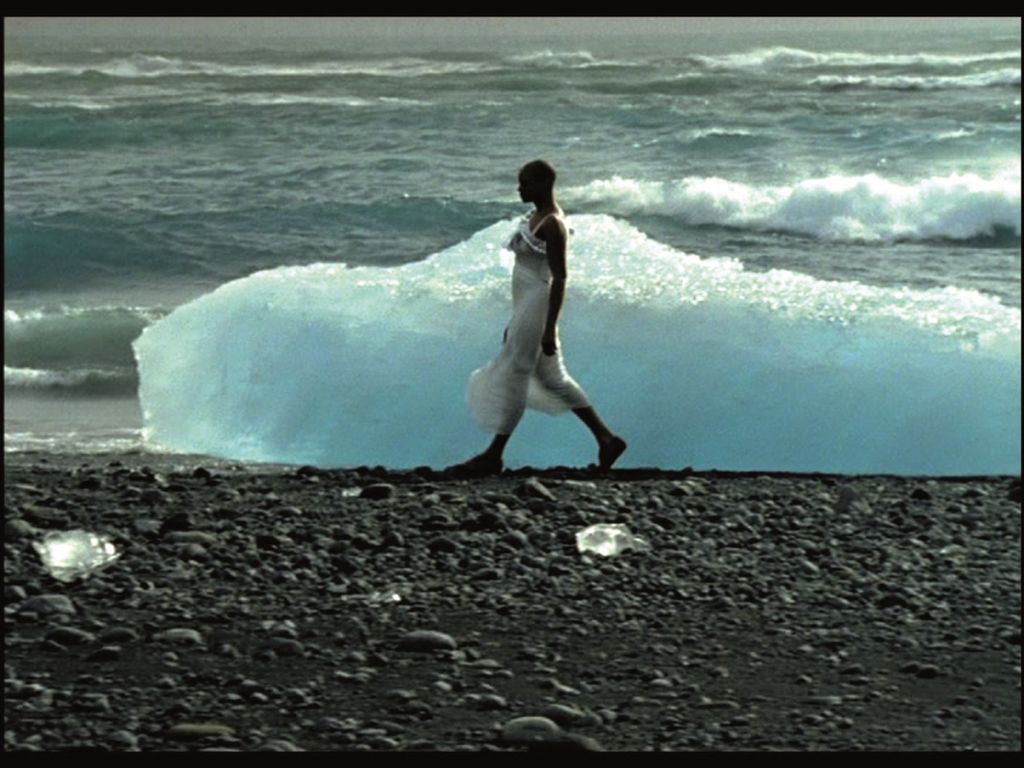 This film still shows a colored, walking woman in profile, in front of an ice floe, behind it the waves break on the beach. Isaac Julien, Sammlung Goetz Munich
