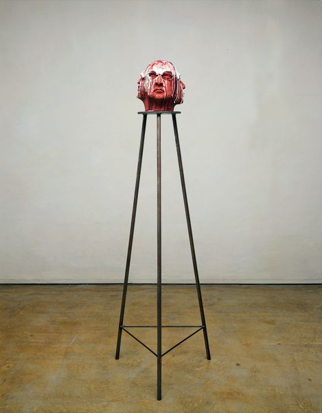 Here you can see a clay head with three faces, over which a glaze of several colours has been poured. The sculpture is placed on a high base with thin legs of black steel.