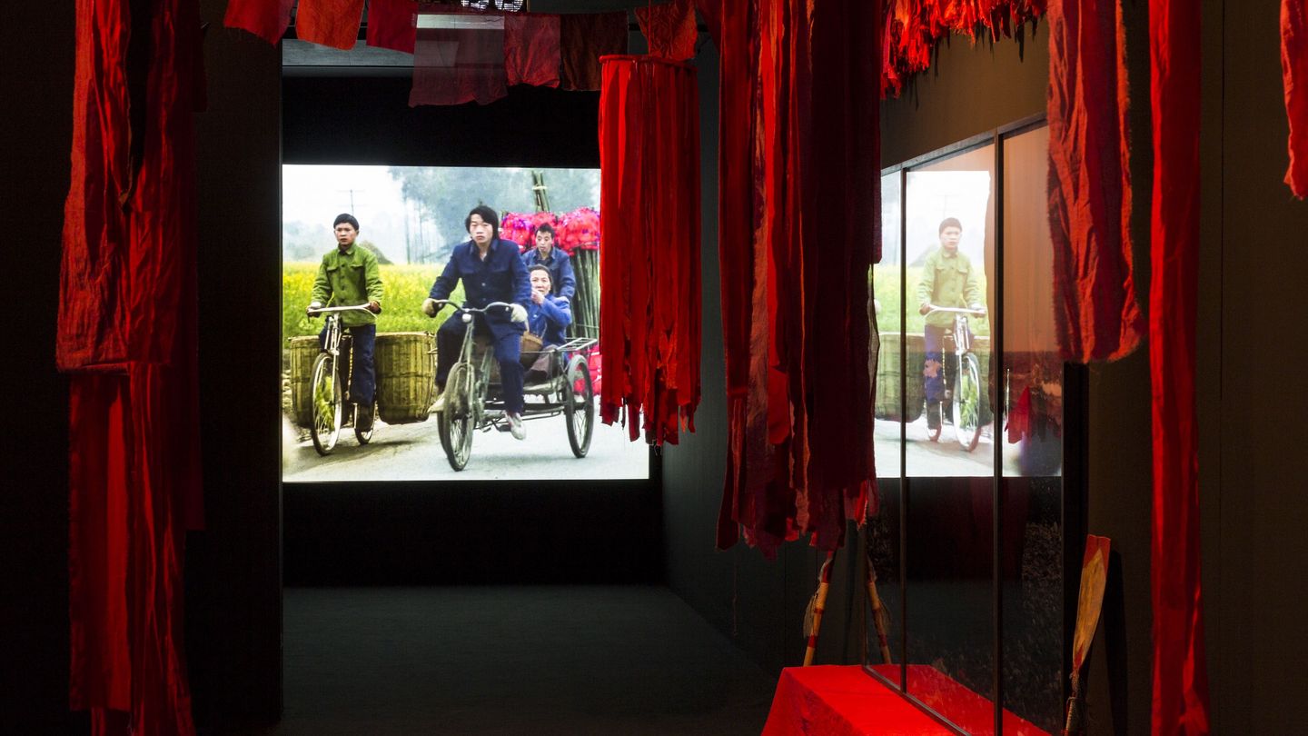 Detail der Installation der Arbeit "Floating Food"; zu sehen sind von der Decke hängende rote Stoffbahnen in einem dunklen Raum sowie ein Video Still, das junge asiatische Männer in traditioneller Arbeitskleidung auf Fahrrädern auf einer Landstraße zwischen grünen Feldern zeigt. Ulrike Ottinger, Sammlung Goetz München 