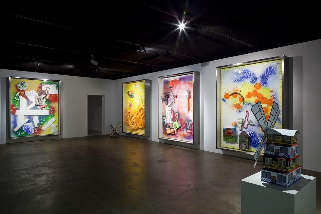 Exhibition view depicting four large, colorful and square paintings in a heavy silver frame. On the floor in one corner and on a pedestal are miniature editions of windmills. Andreas Slominski, Sammlung Goetz Munich 