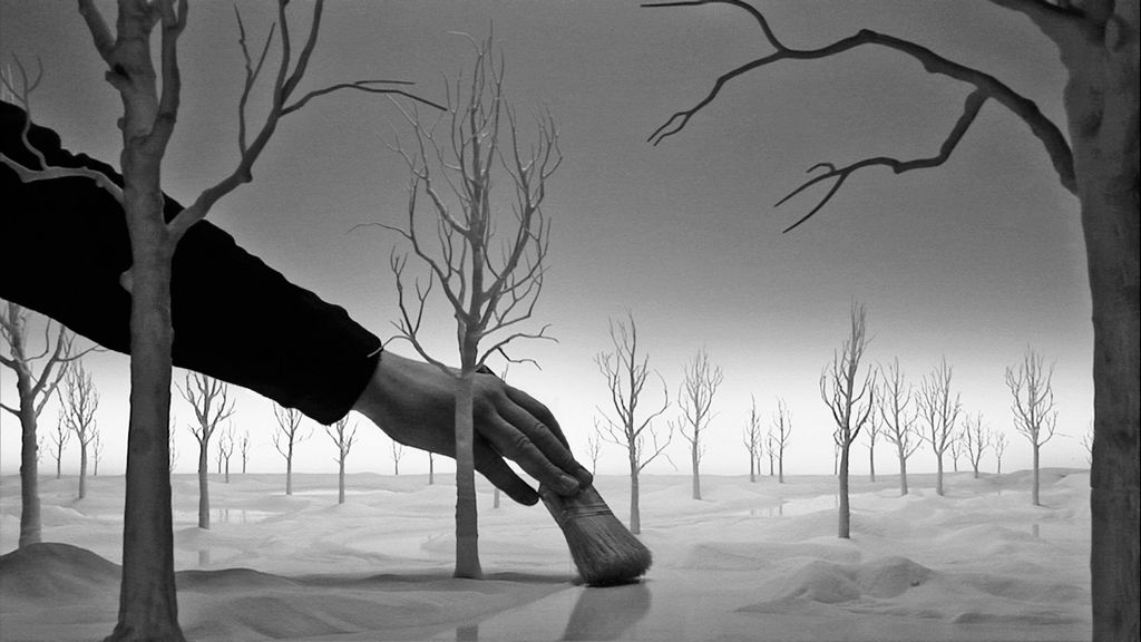 Video Still in black and white showing a model landscape with a river and parched trees, in between is a human hand that appears to be stroking the ground with a brush. Hans Op de Beeck, Sammlung Goetz Munich