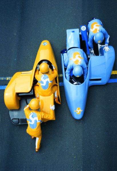 Auf der Fotografie sind zwei Rennwagen nebeneinander in gegenüberliegender Fahrtrichtung zu sehen, das eine ist orange-gelb, das andere himmelblau. Es befindet sich jeweils ein Fahrer in einem gleichfarbigen Einteiler, dahinter stehen Ingenieure in ebenfalls gleichfarbigem Anzug. Matthew Barney, Sammlung Goetz München