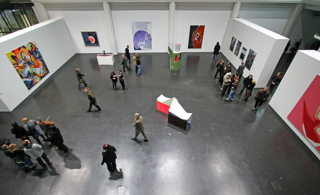Bird's-eye view of the exhibition; white partitions have been drawn into the room, on which paintings and photographs are hung; in the center are visitors and sculptures. Sammlung Goetz Munich