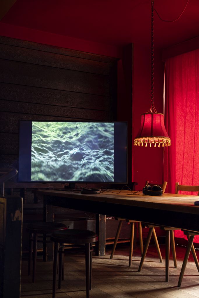 Auf einem Holztisch steht ein Monitor, auf dem eine Art dunkelblaue unregelmäßige Oberfläche gezeigt wird. Sie erinnert an ein Gewässer oder eine Welle. Hiter dem Tisch sind rote Vorhänge zugezogen. Über dem Tisch hängt eine Lampe mit einem altmodischen roten Lampenschirm 