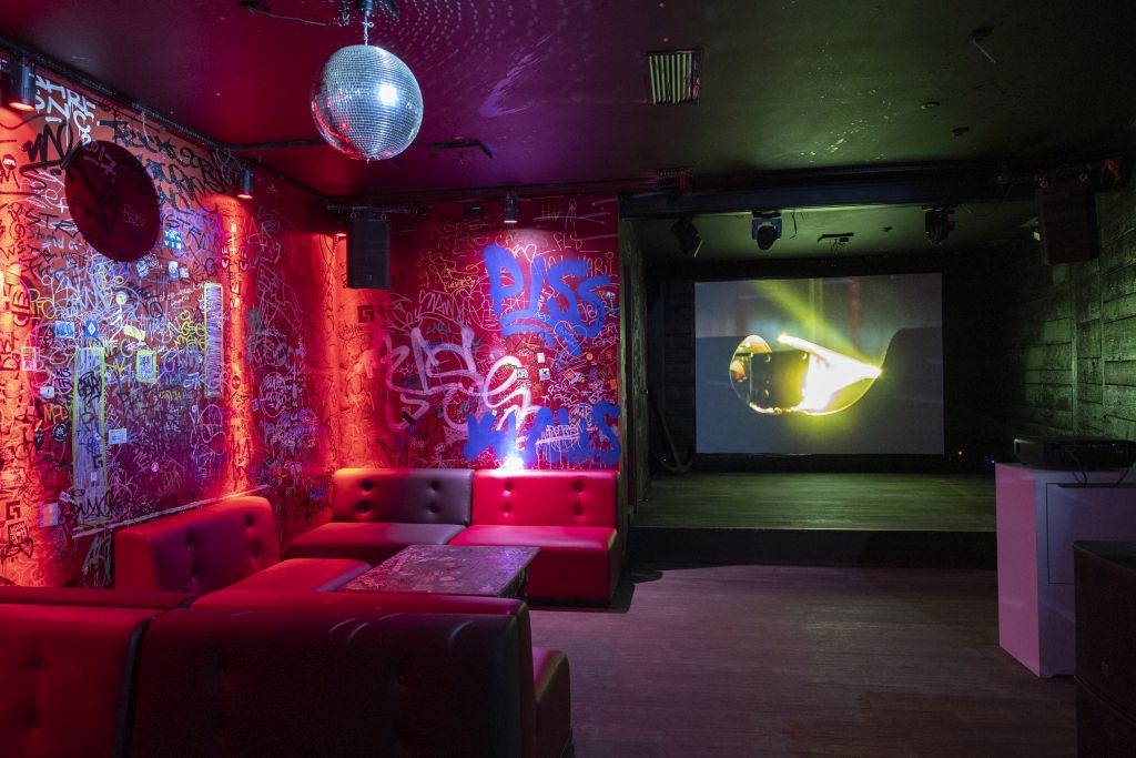Darkened club room with red walls covered in graffiti. On the right is a seating area with red leather sofas over which hangs a disco ball. At the back left is a stage, behind it is a light installation projected on the wall.