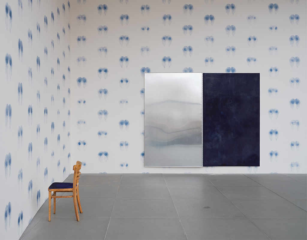 White wallpaper with blue butt prints. On the wall hangs a two-part work reminiscent of a giant ink pad. In front of the wall is a wooden chair with an equally blue seat