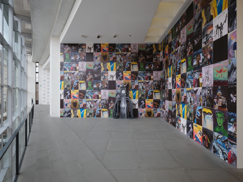 Exhibition room with illustrated wallpaper. The illustrations resemble comic book scenes. In front of the wall is a sculpture with horns, a clock and tentacles. On the left is a window front, in the background you can see the next room