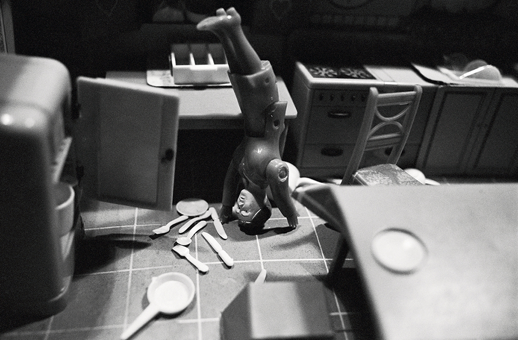Schwarzwießfotografie einer Playmobil-Puppe in einer Puppenküche. Die Puppe macht einen Handstand, neben ihr liegen auf dem Küchenboden Teller und Besteck