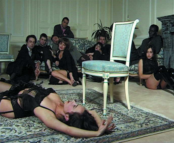 Dieses Videostill zeigt eine Frau in feinen, schwarzen Dessous, die sich auf dem Boden auf einem Teppich räkelt, während sie von einer Personengruppe in einer Ecke des Raumes, bestehend aus Männern in Anzügen und weiteren leicht bekleideten Frauen in schwarz, beobachtet wird
