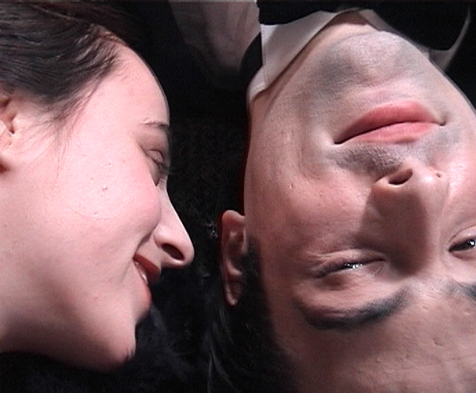 Auf diesem Video Still halten ein Mann und eine Frau ihr Gesichter nebeneinander und lächeln. 