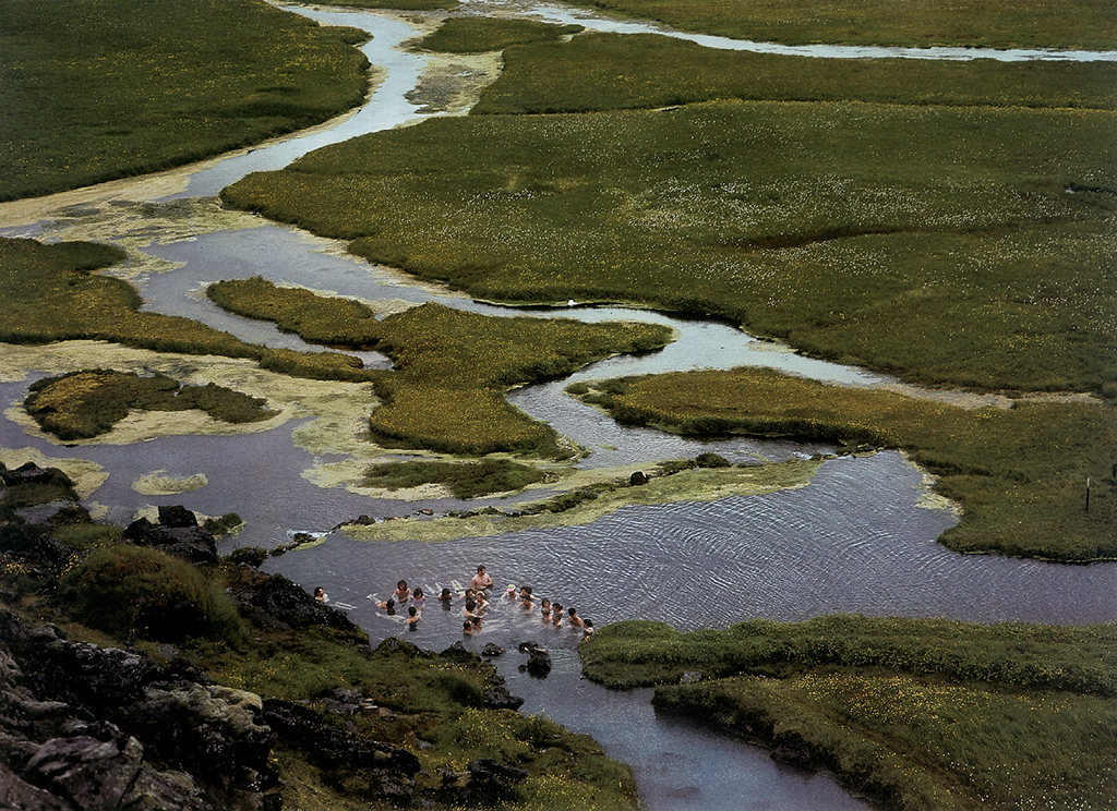 Diese Aufnahme zeigt eine isländische Landschaft mit einer heißen Quelle, in der eine Gruppe von Menschen badet. Die Szenerie ist aus der Vogelperspektive fotografiert worden.