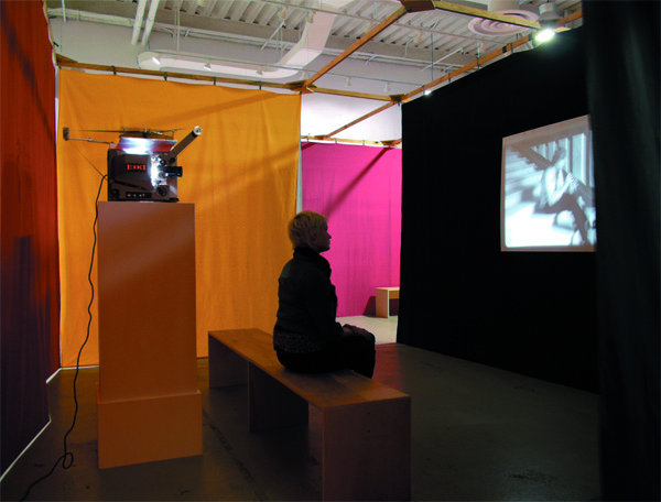 Installation shot showing a visitor sitting on a minimalist wooden bench and watching a video work in black and white. Behind her is the corresponding projector, around her are hanging coloured long fabrics in red, orange and pink. Ulla von Brandenburg, Sammlung Goetz Munich