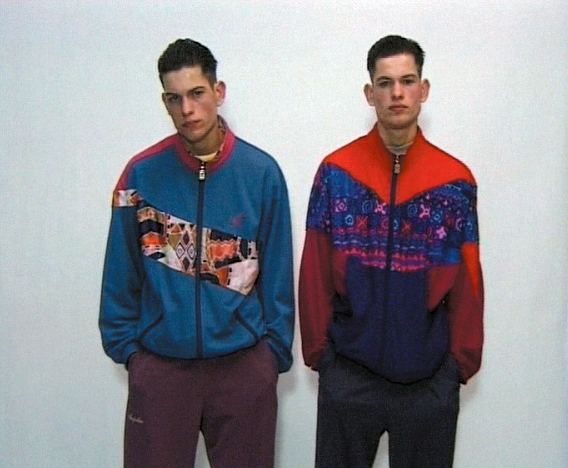 Video Still von zwei jungen Männern mit modischer Kleidung der 1990er Jahre und Händen in den Hosentaschen, die eine coole Haltung suggerieren. Sie stehen vor einer weißen Wand und blicken unbeeindruckt in die Kamera. Rineke Dijkstra, Sammlung Goetz München
