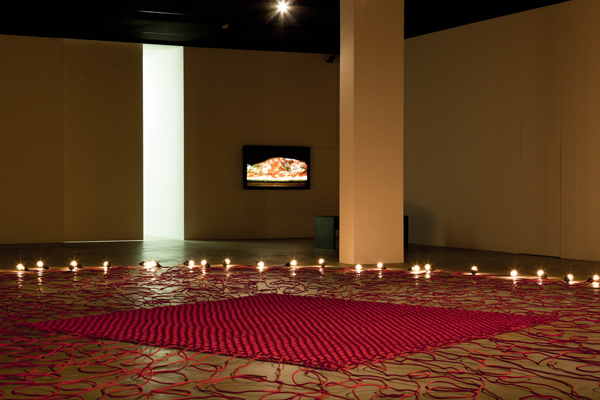 Darkened exhibition space with floor sculpture made of interwoven red electric cables, at the end of each of which is a light bulb that glows. In the background of the picture, a monitor is hanging on the wall on the right, on which a video is running.  