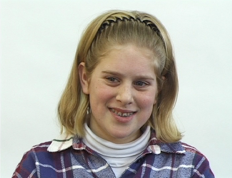Video Still eines Mädchens im Teenager-Alter, mit fester Zahnspange, blondem Haar und kariertem Hemd vor weißem Hintergrund. Rineke Dijkstra, Sammlung Goetz München