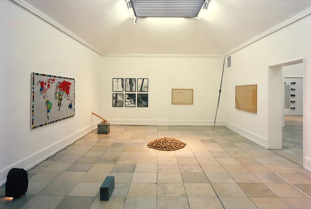This photograph is an installation view with works of the representatives of Arte Povera. A pile of potatoes lies in the middle of the stone floor, on the left side of the wall is an embroidered world map by Alighiero Boetti, and on the wall next to it are six black-and-white photographs hanging upright in two rows of three. Next to it are other two-dimensional works, as well as three-dimensional ones distributed on the floor. 