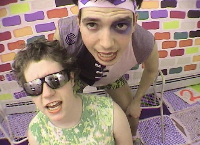 In this video still, two young people can be seen, one wears mirrored sunglasses, the other is made up to look like he has a black eye. Behind them is a colourfully painted wall. Ryan Trecartin, Sammlung Goetz Munich
