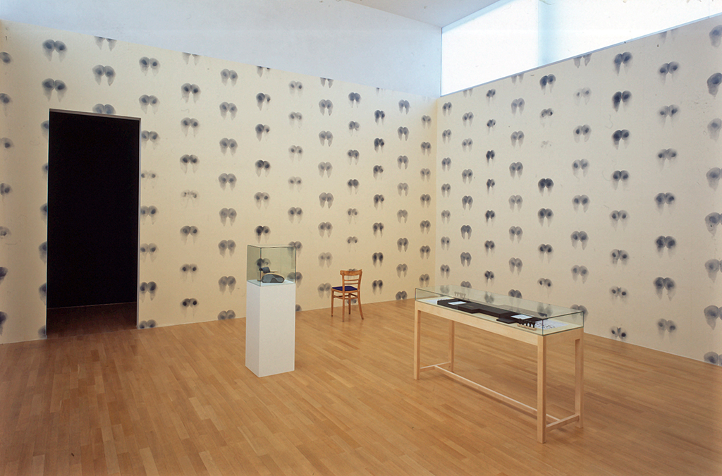 This installation view shows a wallpaper with an even impression of a female breast, an elongated showcase with an object in it, a smaller but higher showcase with object and a wooden chair with dark blue upholstery. All works are by the artist Abigail Lane. 