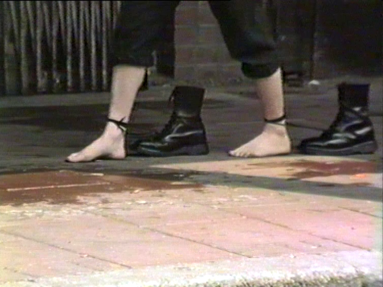 Film still, bare feet stride across a pavement. Black Dr. Martens boots are attached to their ankles with shoelaces, which they drag behind them step by step.