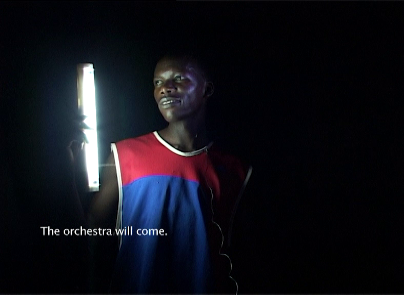 Video Still, a young black man stands in the darkness with a neon tube in his hand, which illuminates him from the side. His face is turned towards the lamp, he looks past it and smiles. In the lower left of the image it says "The orchestra will come."
