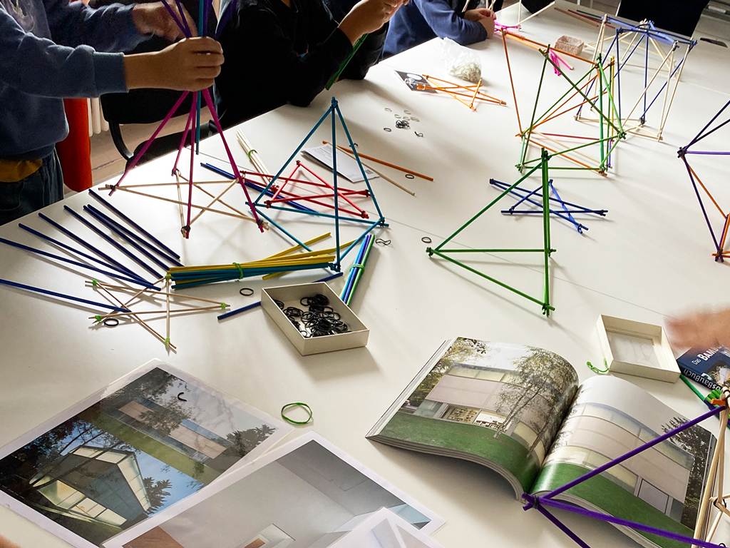 Auf einem Tisch liegen Architekturbilder der Sammlung Goetz, daneben bauen Kinder räumliche Strukturen aus bunten Strohhalmen