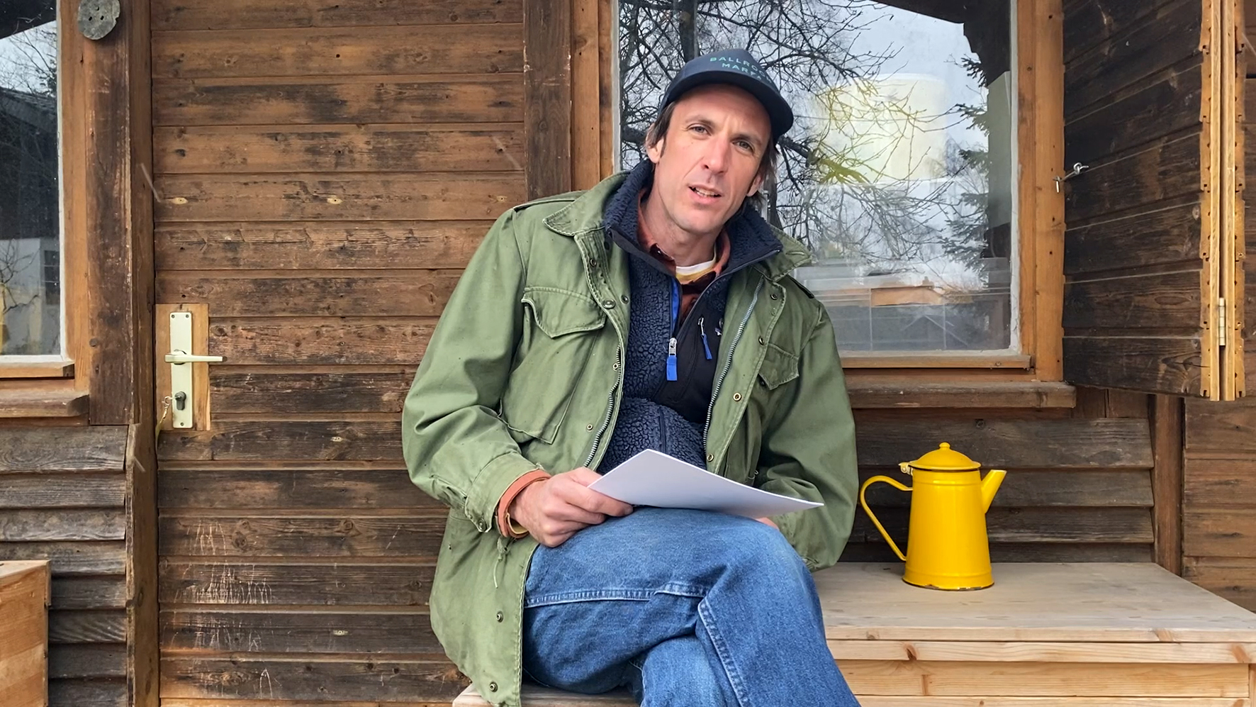 Cyrill Lachauer sitzt im Freien vor einer Holzhütte, neben ihm steht eine gelbe emaillierte Teekanne. Er hält einen Stapel Papierblätter in der Hand und Blicht in Richtung des Betrachters, der Mund halboffen, als würde er gerade sprechen. Sammlung Goetz, München