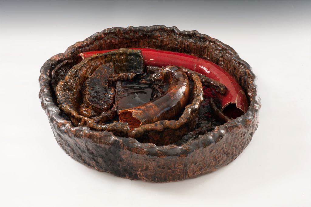 A large clay bowl is filled with sausage-shaped ceramic elements. The colouring shifts between brown and red. The surfaces display an irregular pattern, and the glaze is highly glossy.