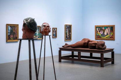 Ausstellungsaal mit hellblau gestrichenen Wänden mit Skulpturen von Thomas Schütte (Bemalte Tonköpfe auf hohen Sockeln und große Stahlskulptur einer liegenden Frau) und Gemälden von May Beckmann