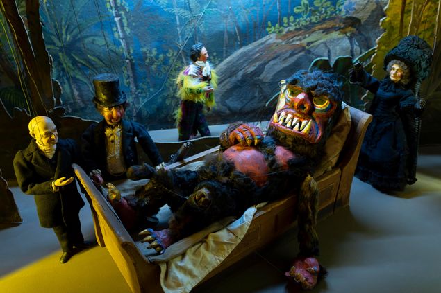 Marionetten auf einer Bühne: Ein haariges Monster mit großen Zähnen liegt in einem Bett. Drumrum stehen vier menschliche Figuren