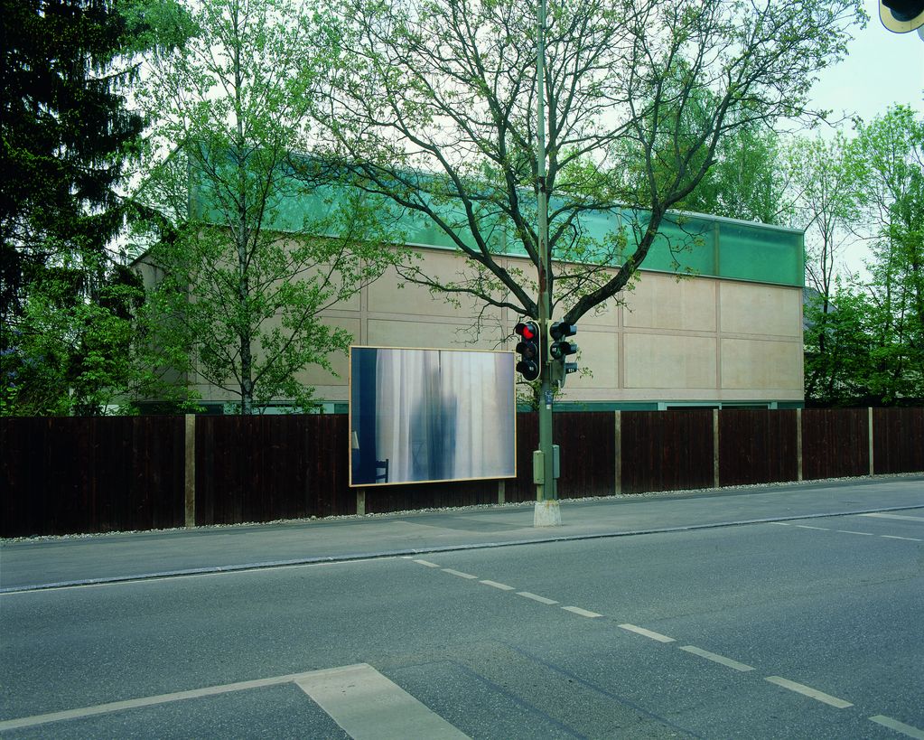 The picture shows an exterior view of the exhibition building as seen from the street. A large billboard is attached to the continuous dark brown fence, showing a photographic work by Felix Gonzalez-Torres. It depicts a white, translucent curtain with a person behind it casting a slight shadow.