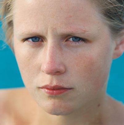 © Roni Horn Colour photograph, portrait of a young blond, blue-eyed woman. Her shoulders are bare and water is dripping from her face. She is standing in front of a radiant light blue background. Roni Horn, Goetz Collection, Munich