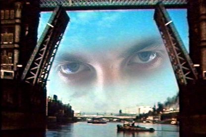 This film still shows two superimposed shots of London Tower Bridge, which has opened its flaps to allow larger ships to pass through. In the background, there is actually a smaller steamboat from which black smoke is coming out. In the upper part of the picture, there is in turn a pair of eyes that look intensively into the camera and are flanked by the open locks.